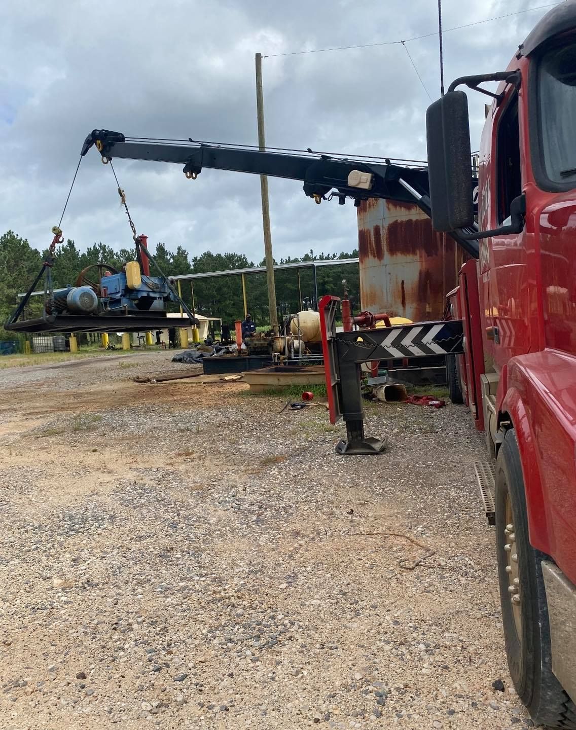 Red truck with crane lifting a flatbed trailer in a gravel yard.