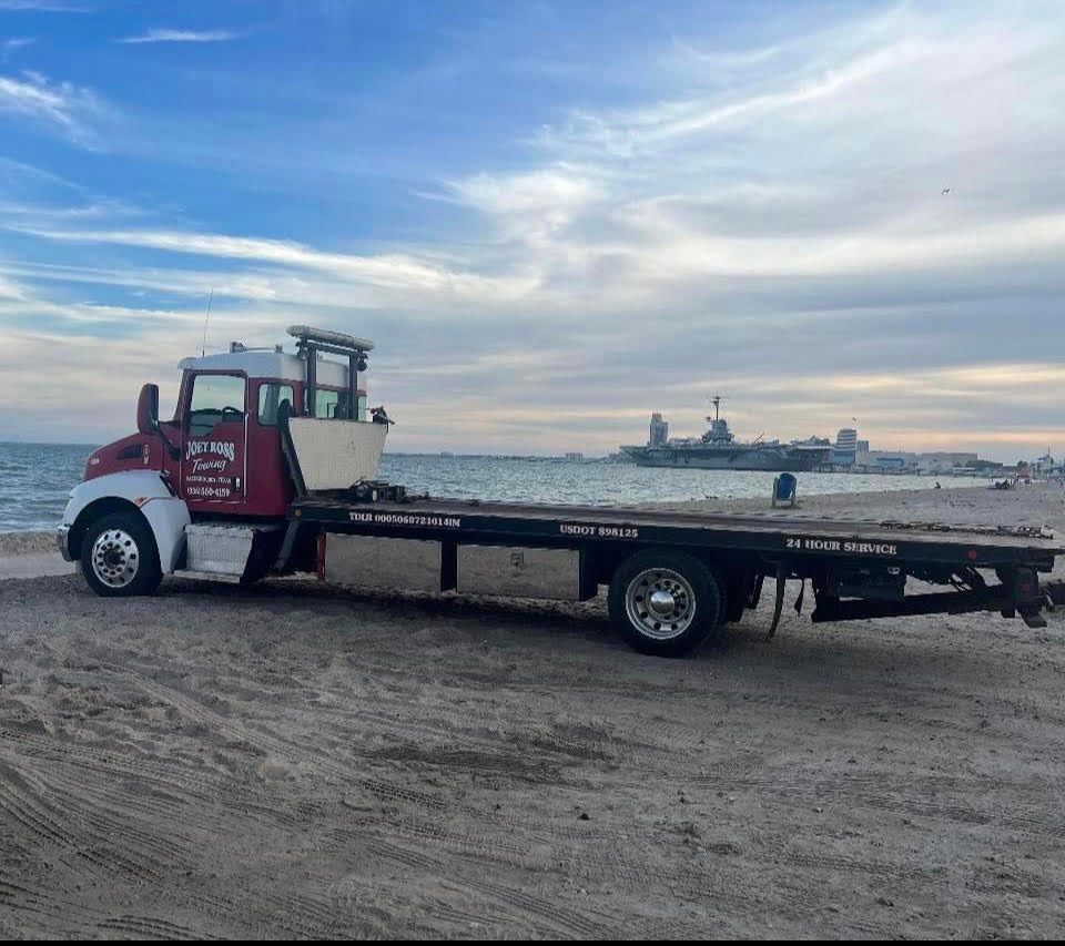 Red flatbed tow truck on a sandy beach with ships on the horizon at sunset