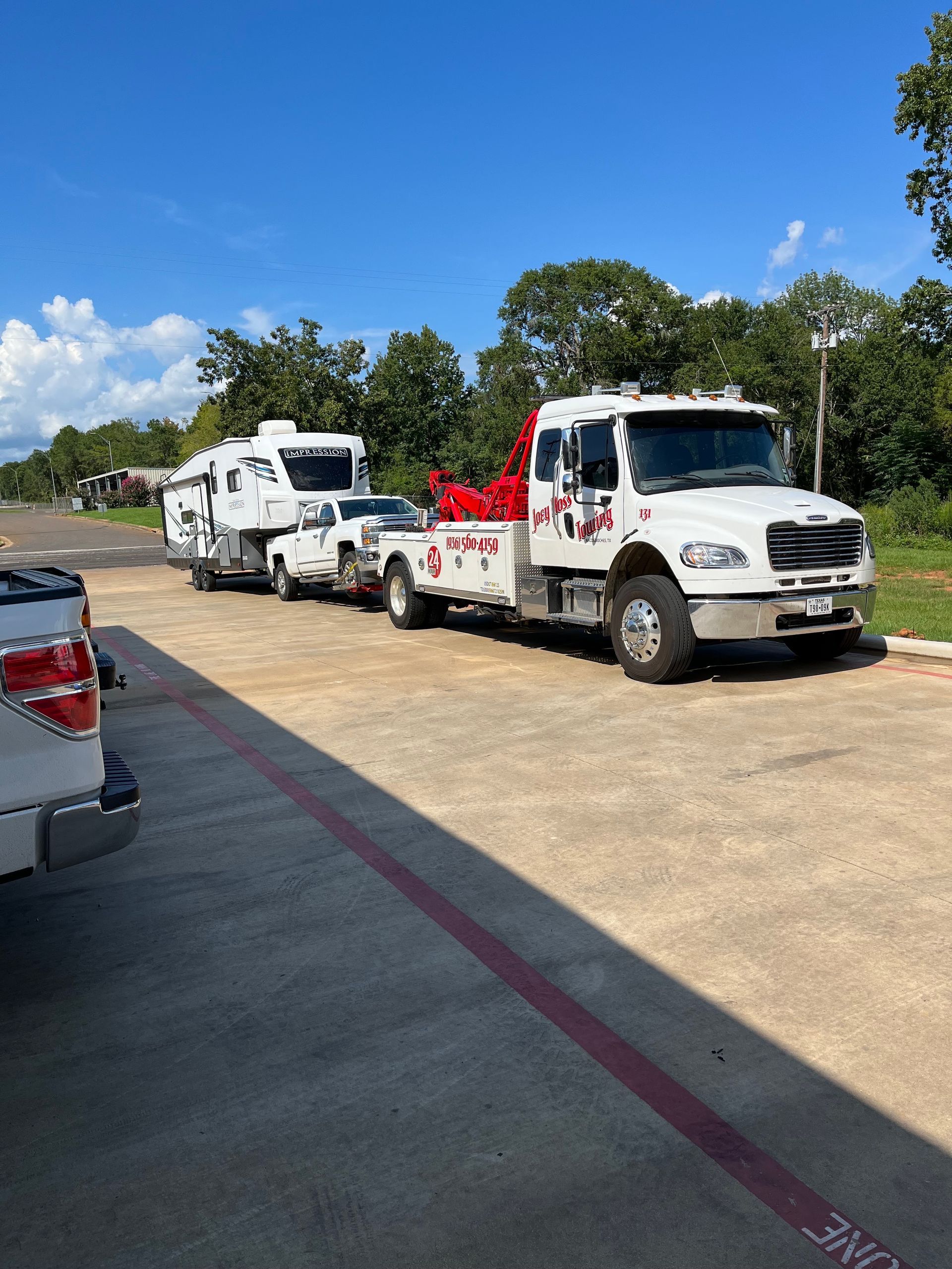 Tow trucks parked along a roadside under a blue sky, one white and one red, with trees in the background