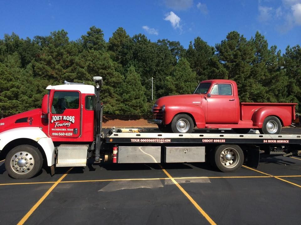 Red flatbed tow truck carrying a red pickup in a parking lot with trees in the background