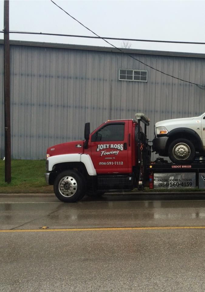 Red tow truck hauling a white SUV on a rainy street beside a gray warehouse.