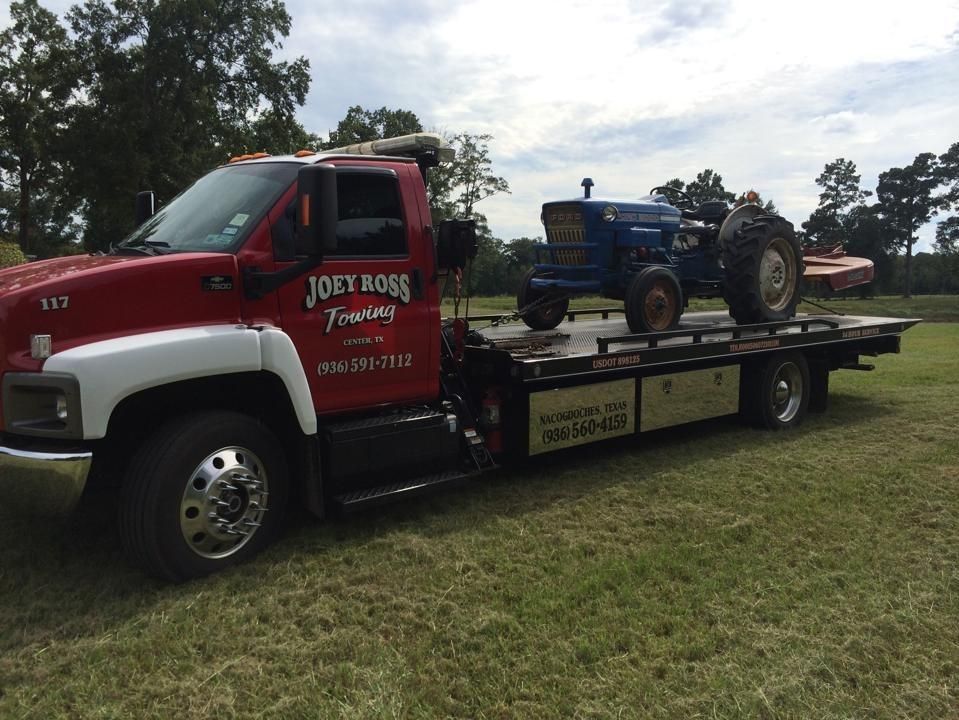 Red tow truck carrying a blue tractor on a flatbed in a grassy field