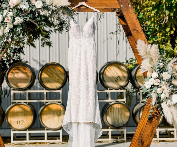 White wedding dress hanging on a wooden arch, with wine barrels and greenery in the background.