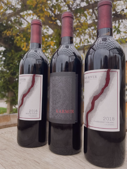 Three red wine bottles with labels on a stone surface outdoors, with blurred trees in the background