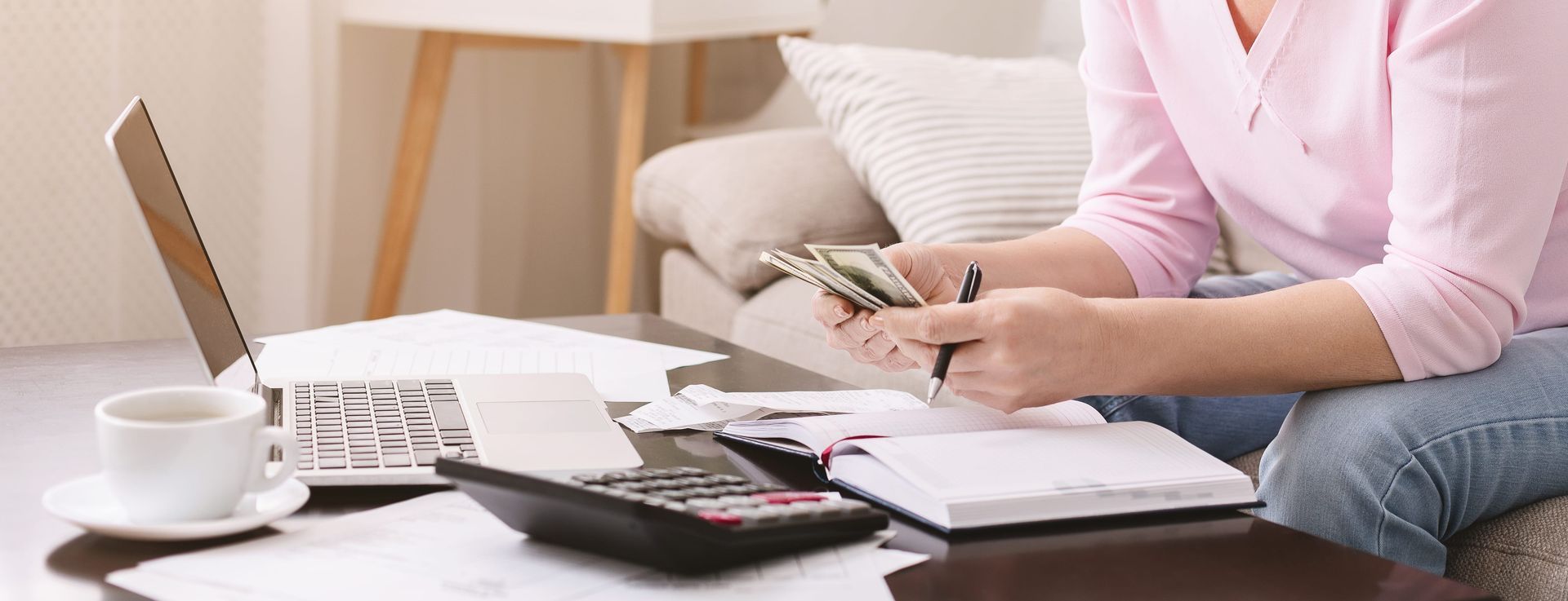 A person in a pink shirt sits on a sofa, counting cash while managing personal finances with a calculator and notebook.