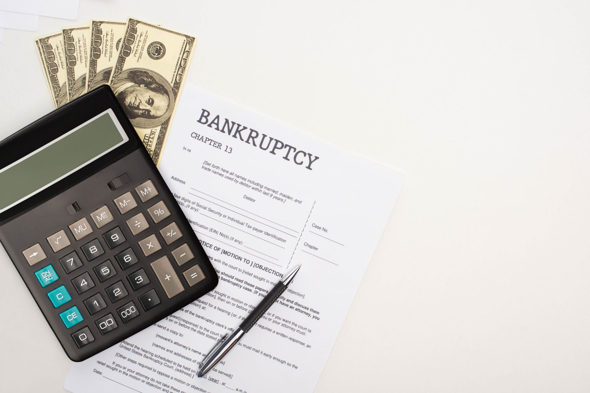 A black calculator, a pen, and three hundred-dollar bills sit atop a document titled Bankruptcy on a white surface.