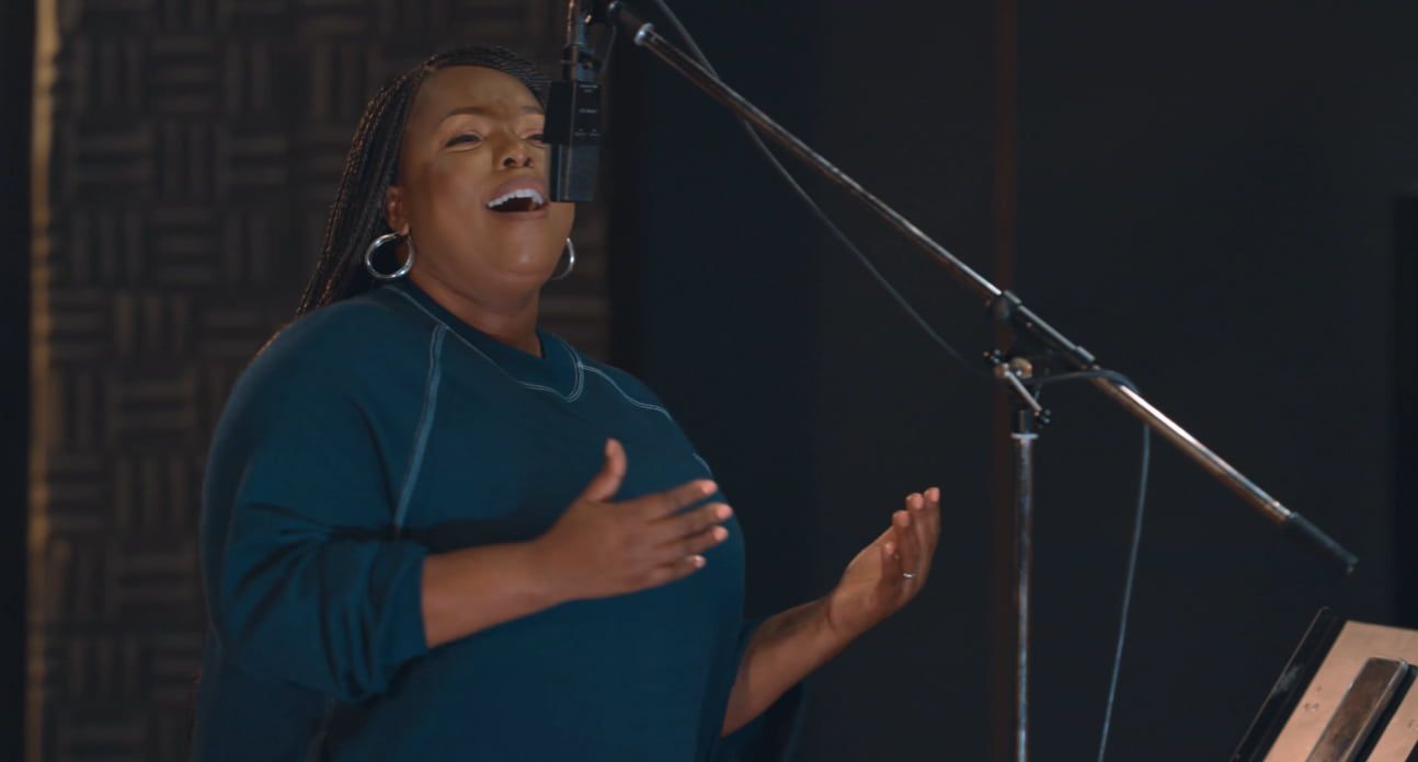 Chevelle singing into a microphone in a recording studio, wearing a teal top, with hands raised.