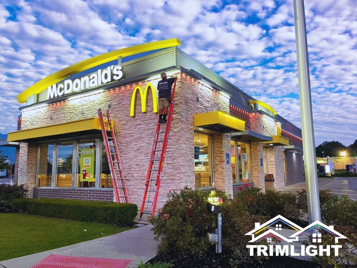 Person on a ladder installing lights on a McDonald's building. The building has a yellow and brick exterior.