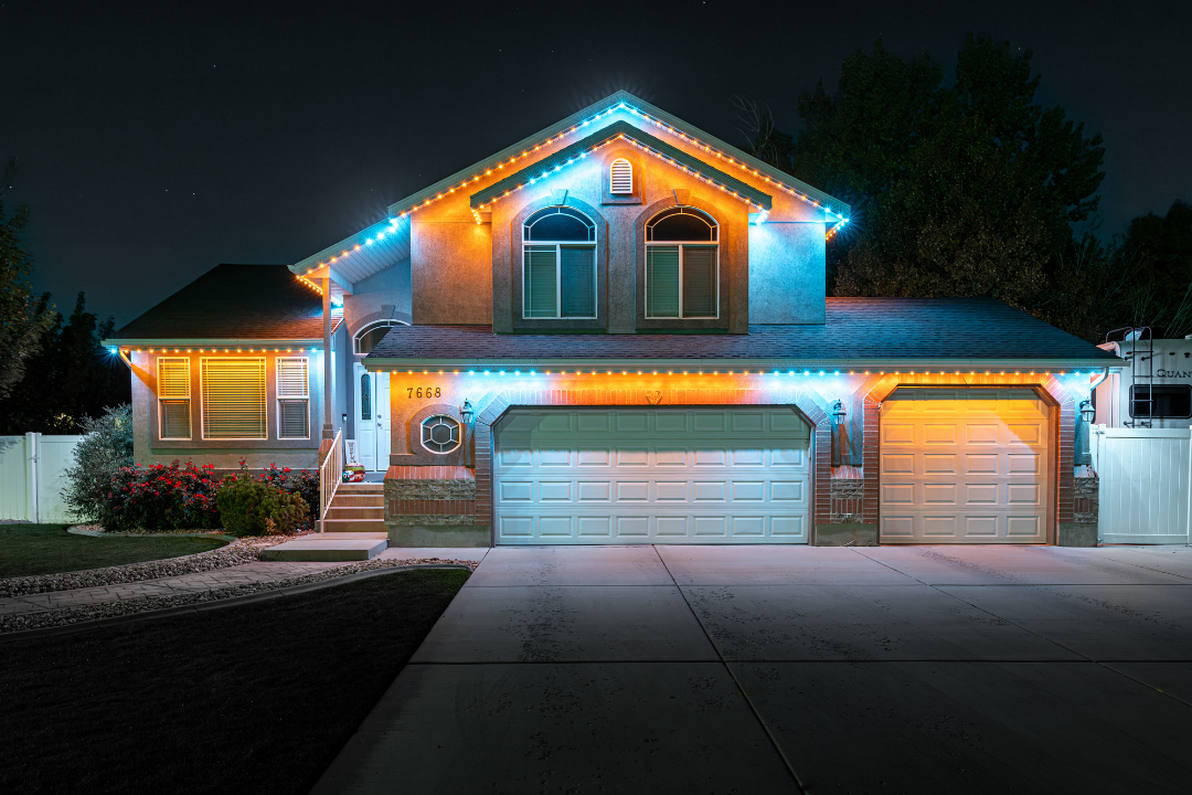House at night lit with blue and orange lights, driveway, garage doors, dark sky.