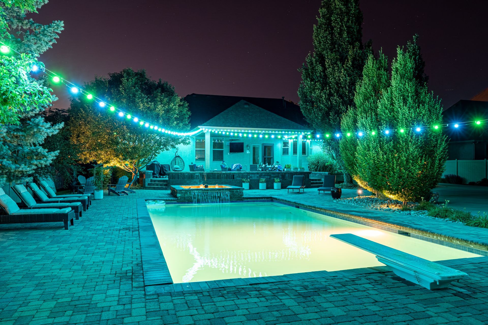 Night view of a backyard pool lit with turquoise lights, string lights, and a fire pit, with a house in the background.