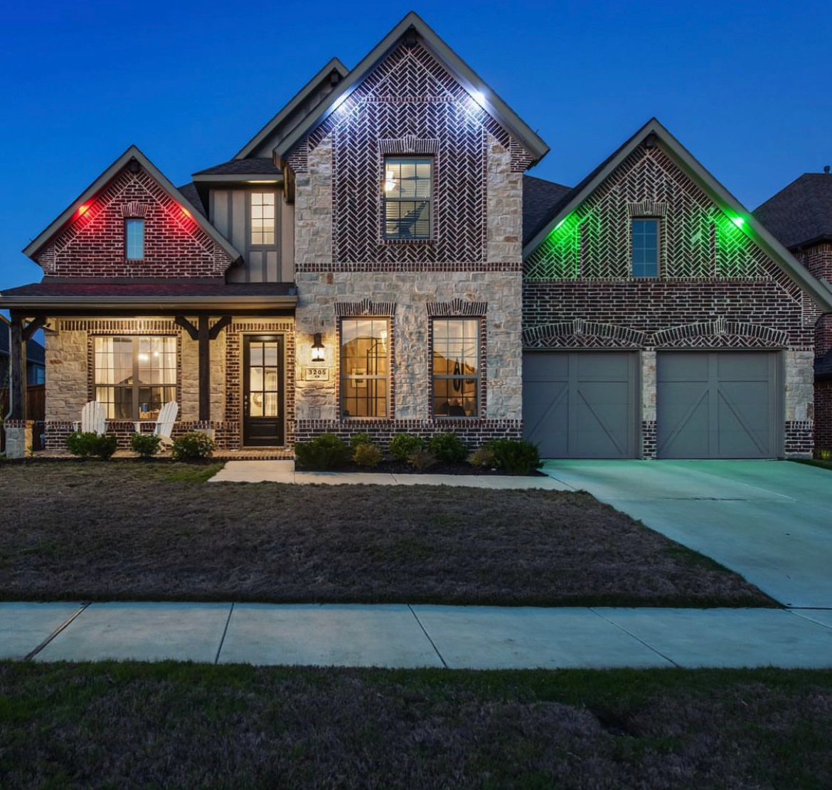 Two-story house with Christmas lights; red above the porch, green above the garage. Evening setting.