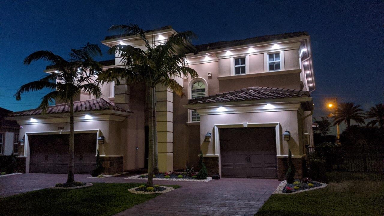 Two-story beige house at night, lit with bright overhead lights. Palm trees and a dark sky surround.