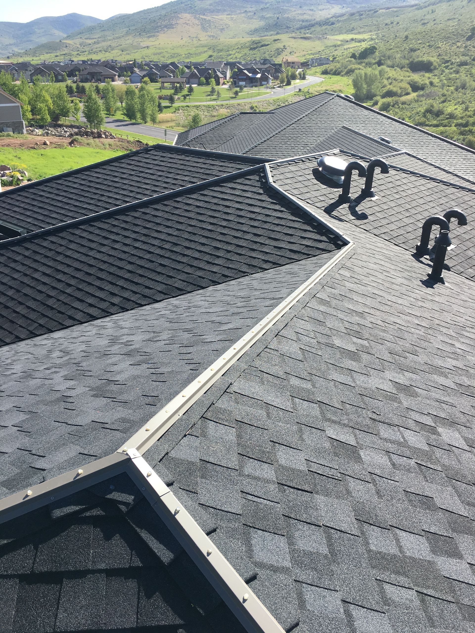 Dark asphalt shingle roof with vents and flashing, overlooking a neighborhood and distant mountains.