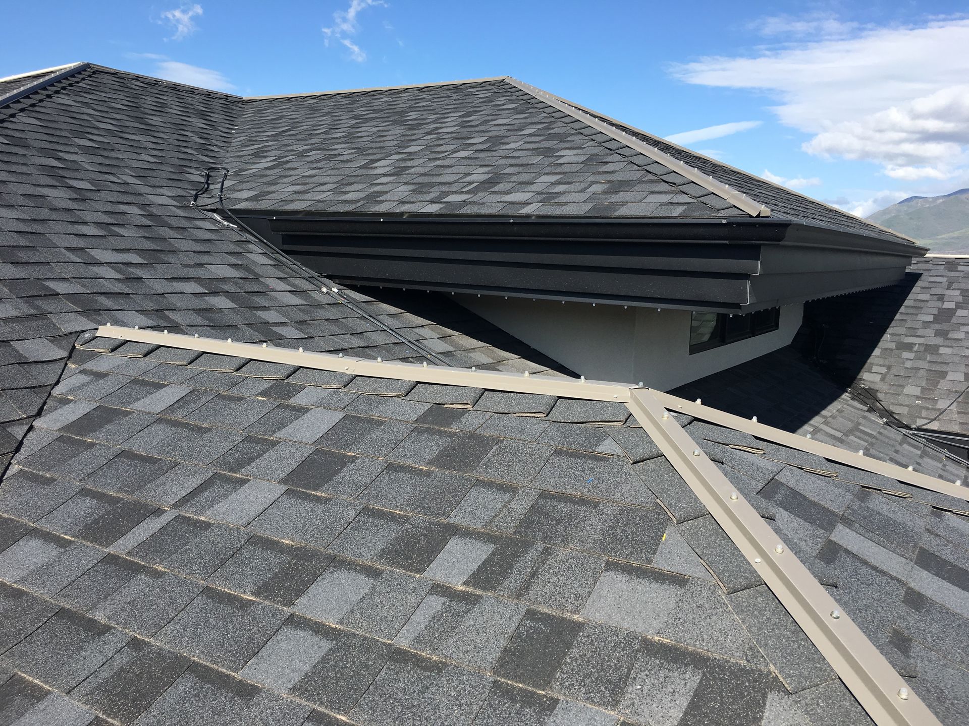 Dark gray asphalt shingle roof on a house with a dormer and light blue sky.