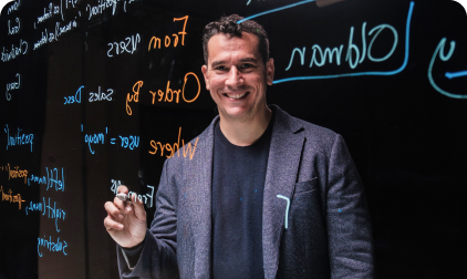 Man smiling, pointing at a whiteboard with colorful handwritten notes.