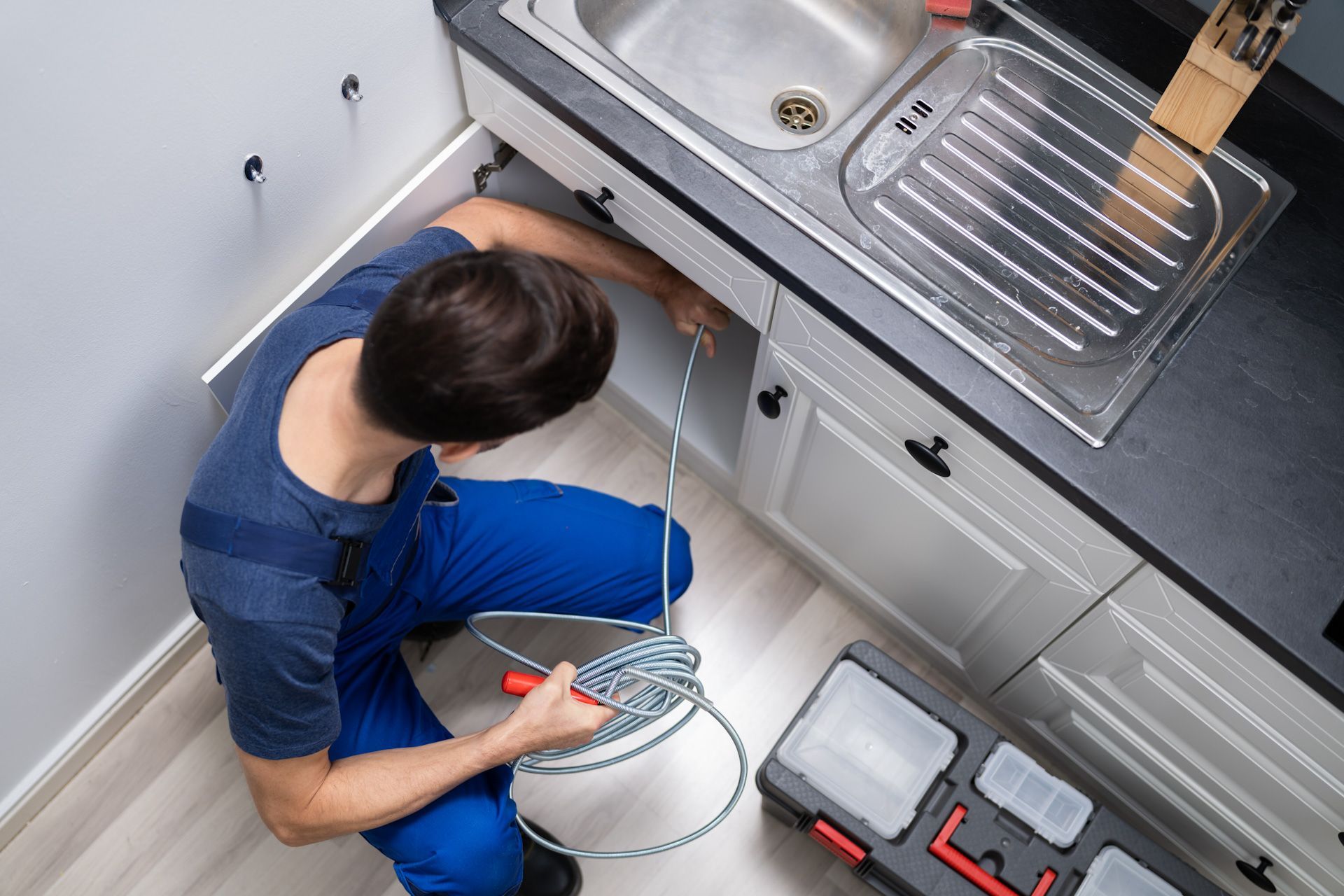 A plumber is kneeling down under a sink in a kitchen.