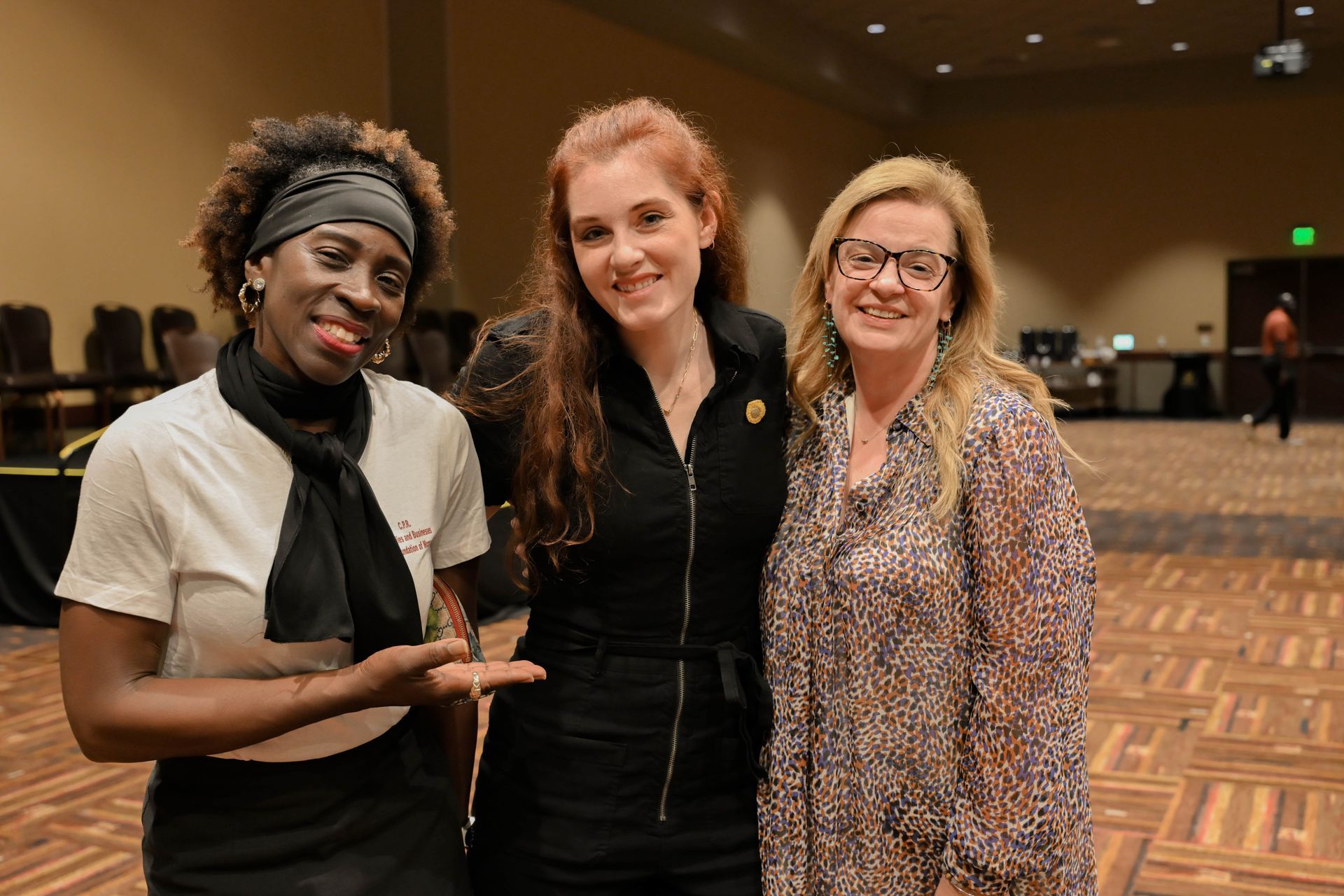 Three people smiling, posing together indoors. Woman on left points to her right.