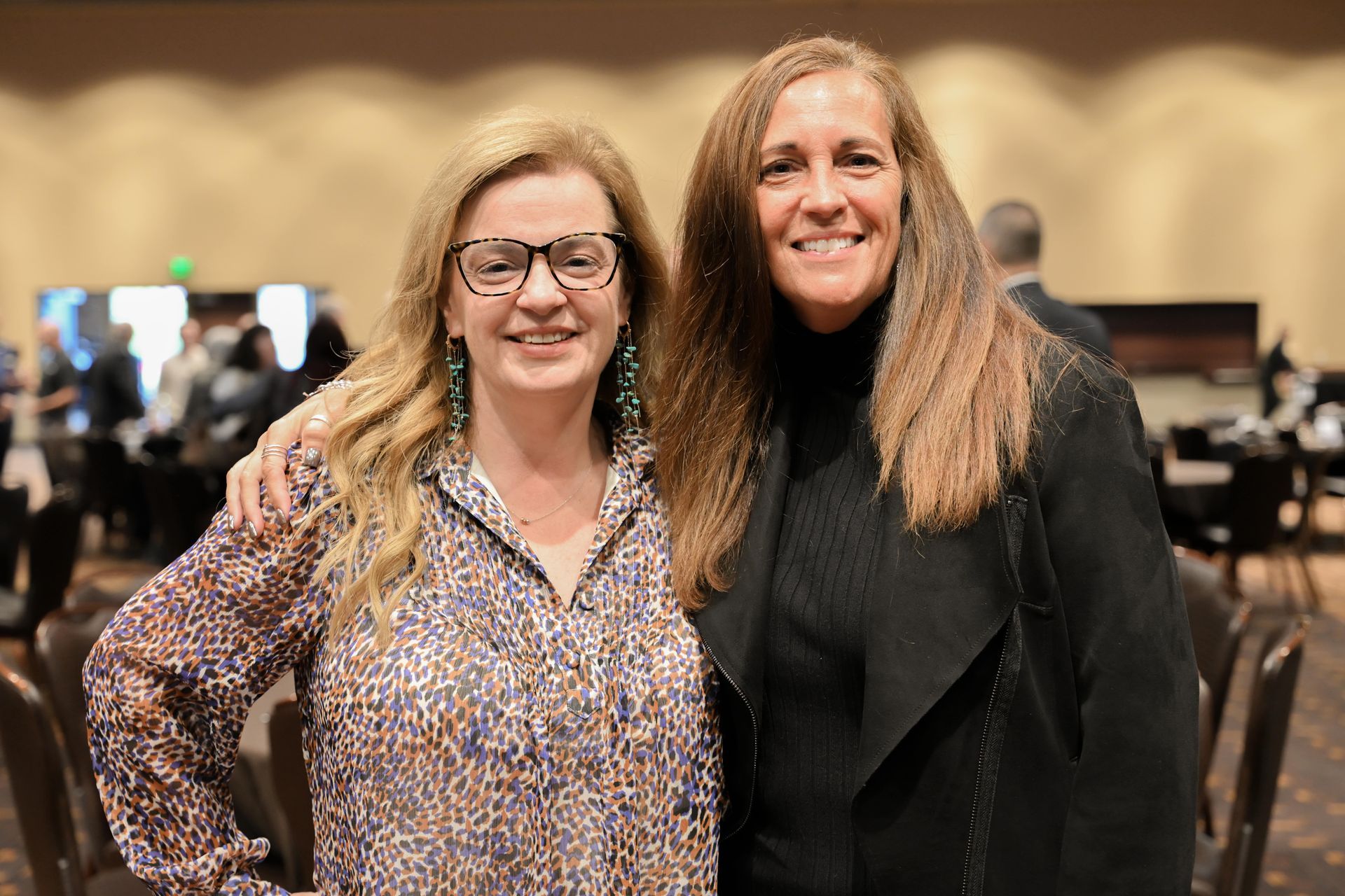 Two women smiling, arm-in-arm. One wears glasses and a patterned top, the other a black jacket. Indoors, blurred background.