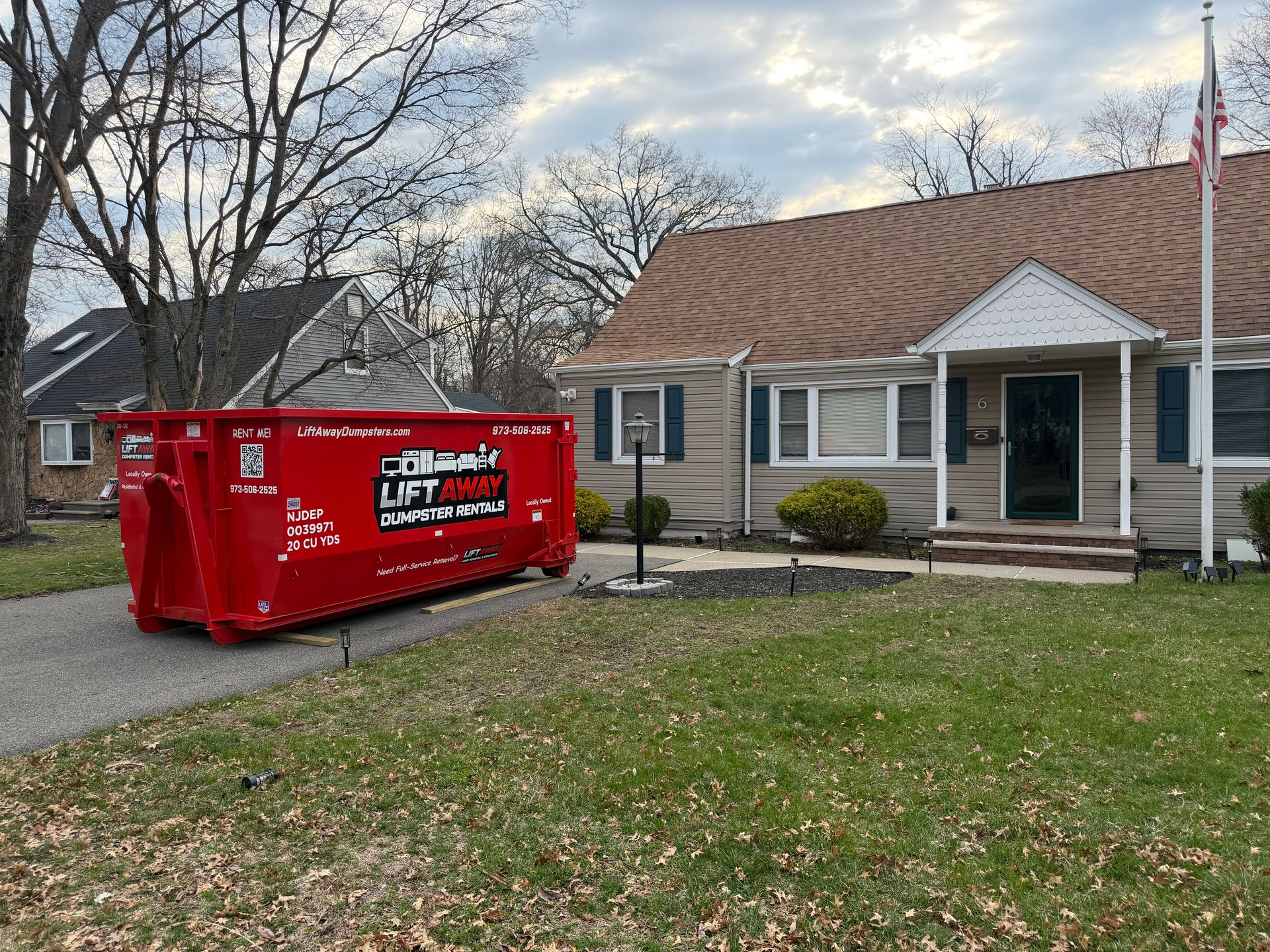 A bright red dumpster sits on the driveway in front of a tan house with brown shingles on a cloudy day.