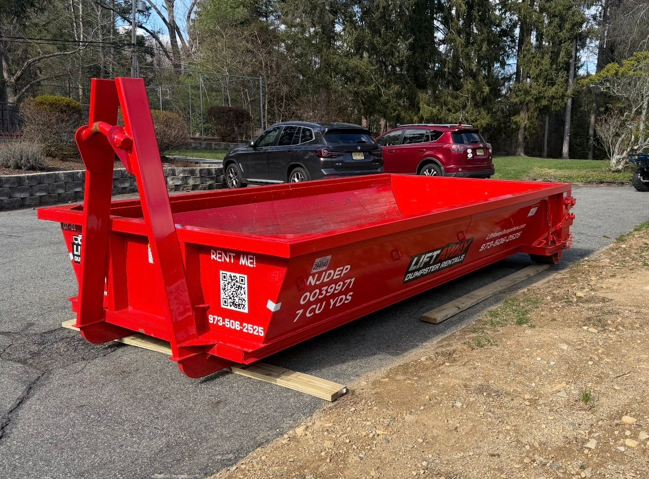 A bright red, low-profile roll-off dumpster sitting on a gravel driveway with cars parked in the background.