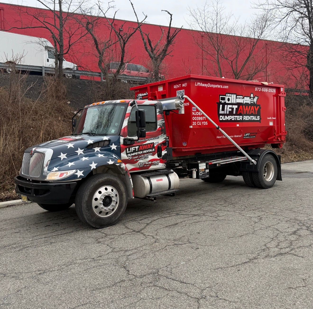 A red dumpster truck with an American flag paint job on the cab, parked on an asphalt lot in front of a red building.