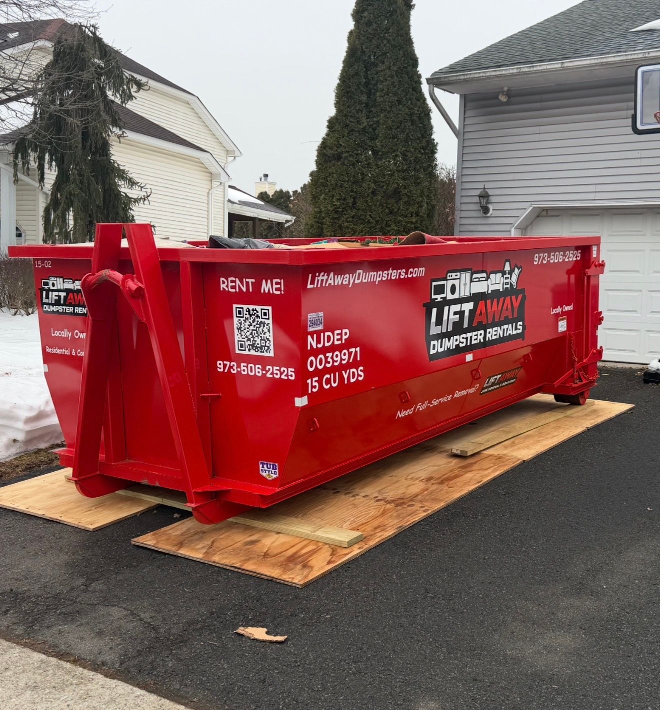 A bright red roll-off dumpster sits on wooden boards in a driveway in front of a residential home.