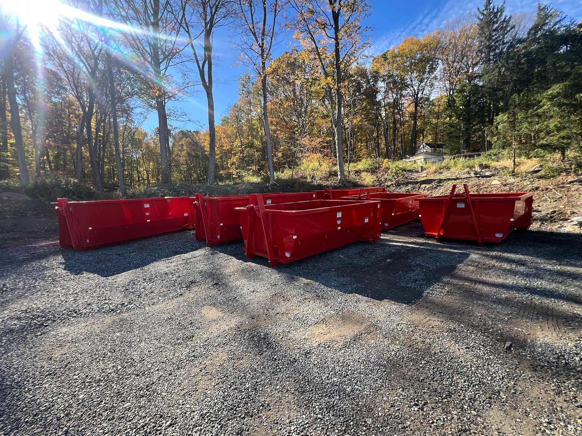Red dumpsters sit on gravel in a wooded area with autumn trees.