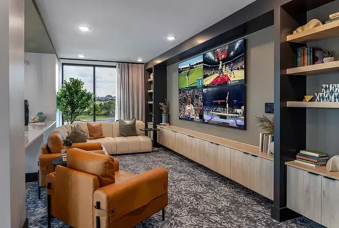 Modern living room with tan chairs, gray sofa, large wall TV, and city view through window
