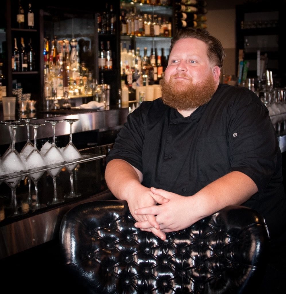 Bartender in a black shirt seated at a bar with bottles and glassware in the background