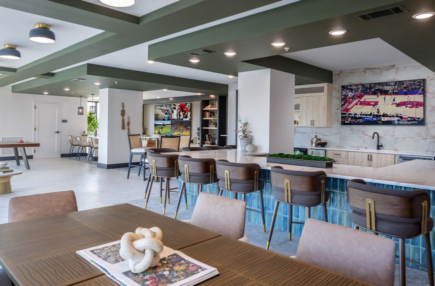 Modern café interior with wooden tables, bar stools, and a long counter under green ceiling beams.