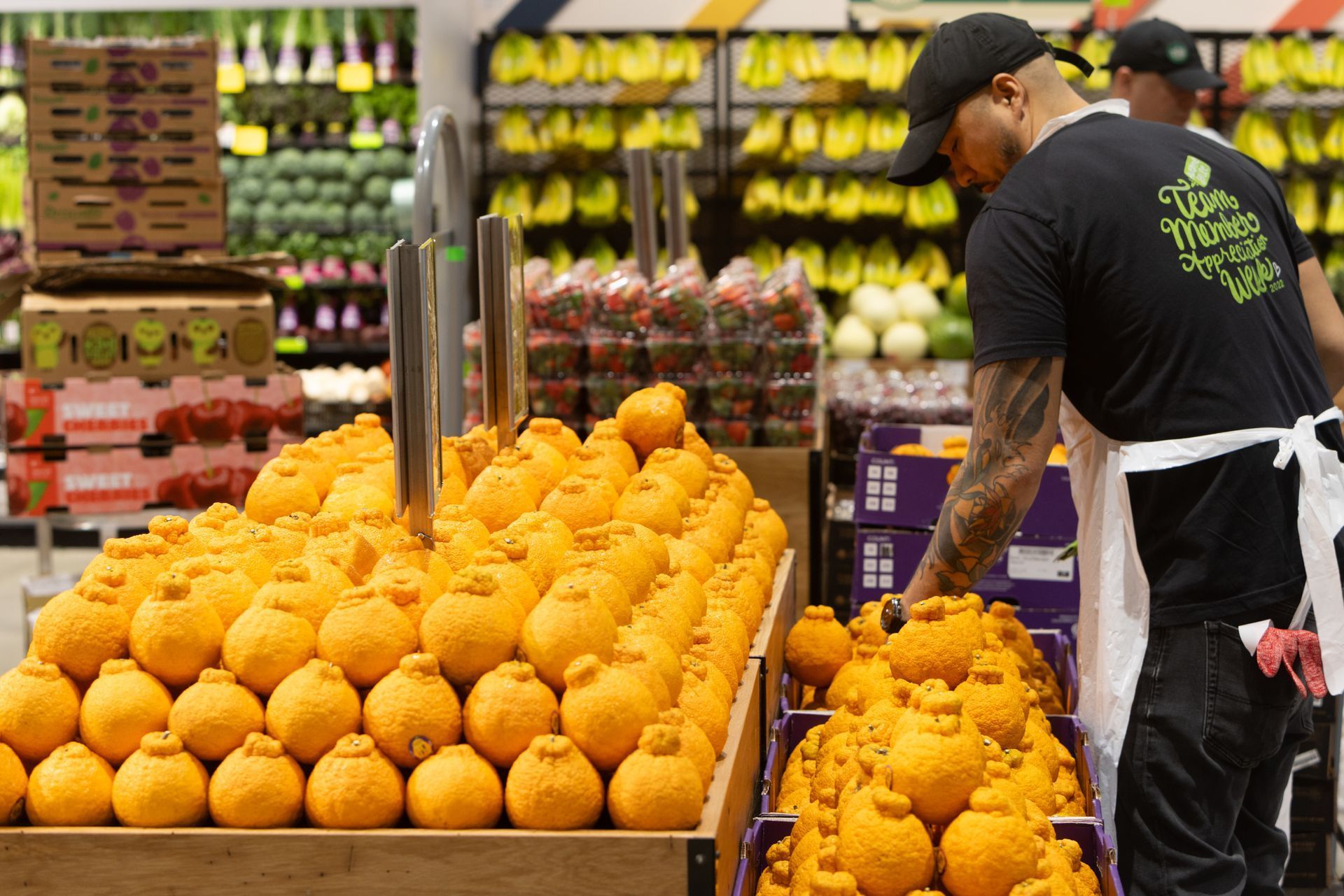 Produce section in grocery store with oranges and bananas; worker in black shirt arranging fruit.