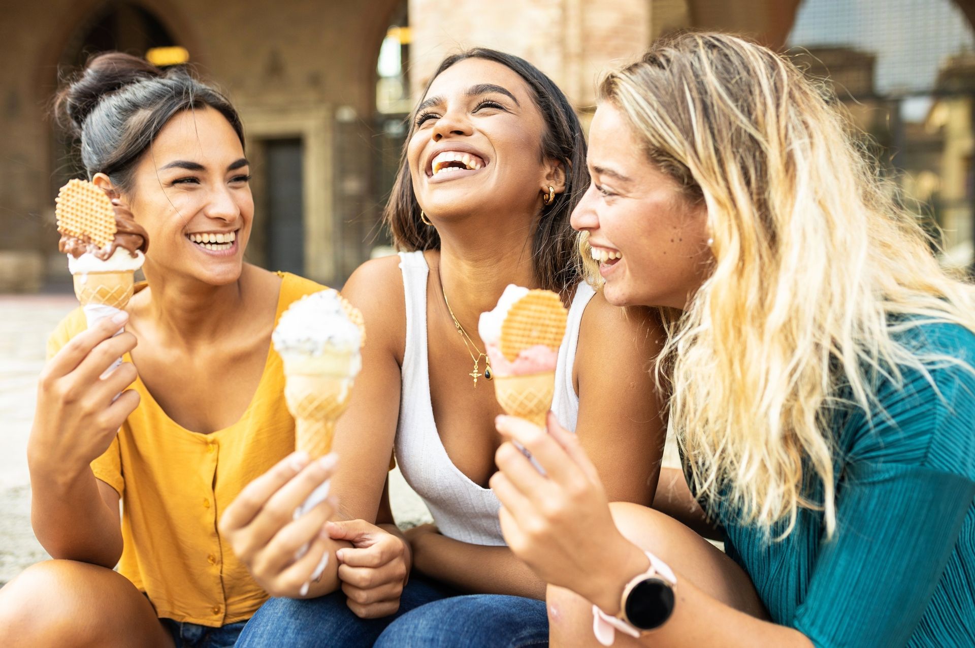 Three friends laughing and eating ice cream cones outdoors at a table