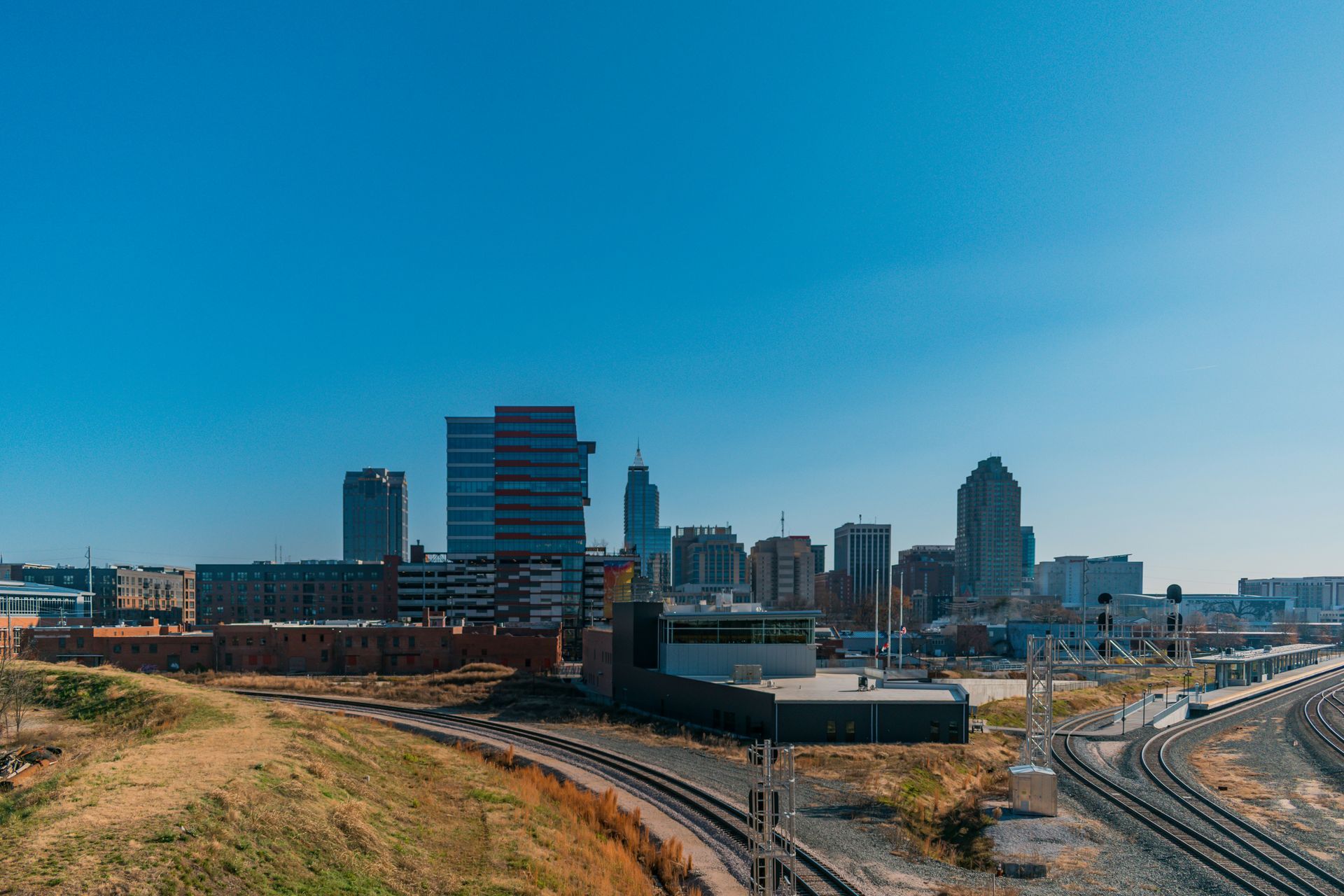 A bright, sunny view of a city skyline with modern buildings, train tracks in the foreground, and a clear blue sky.