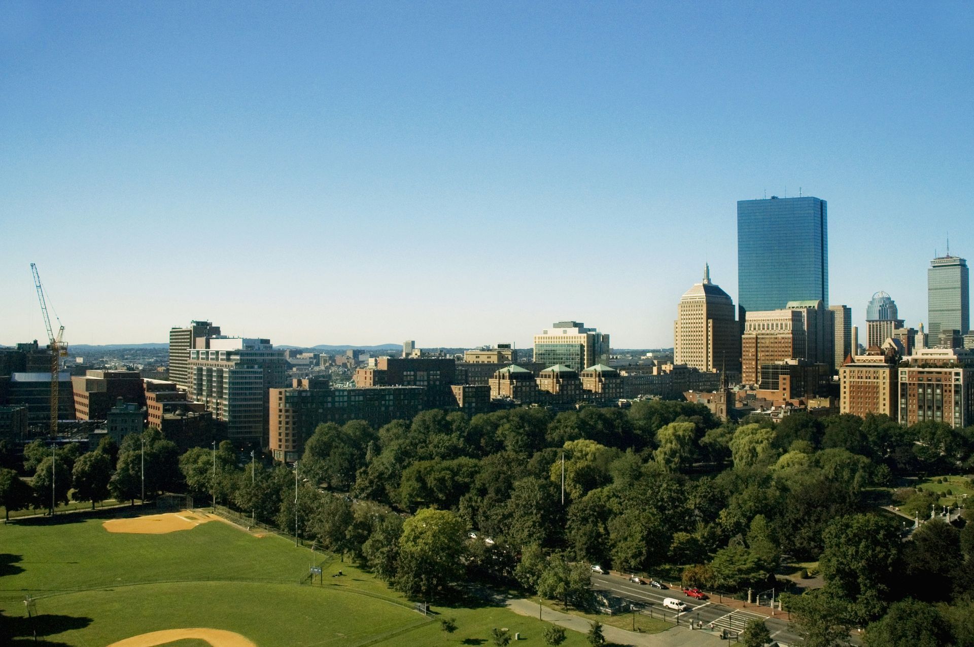 A panoramic view of the Boston skyline with green trees, a baseball diamond, and tall buildings under a clear blue sky.