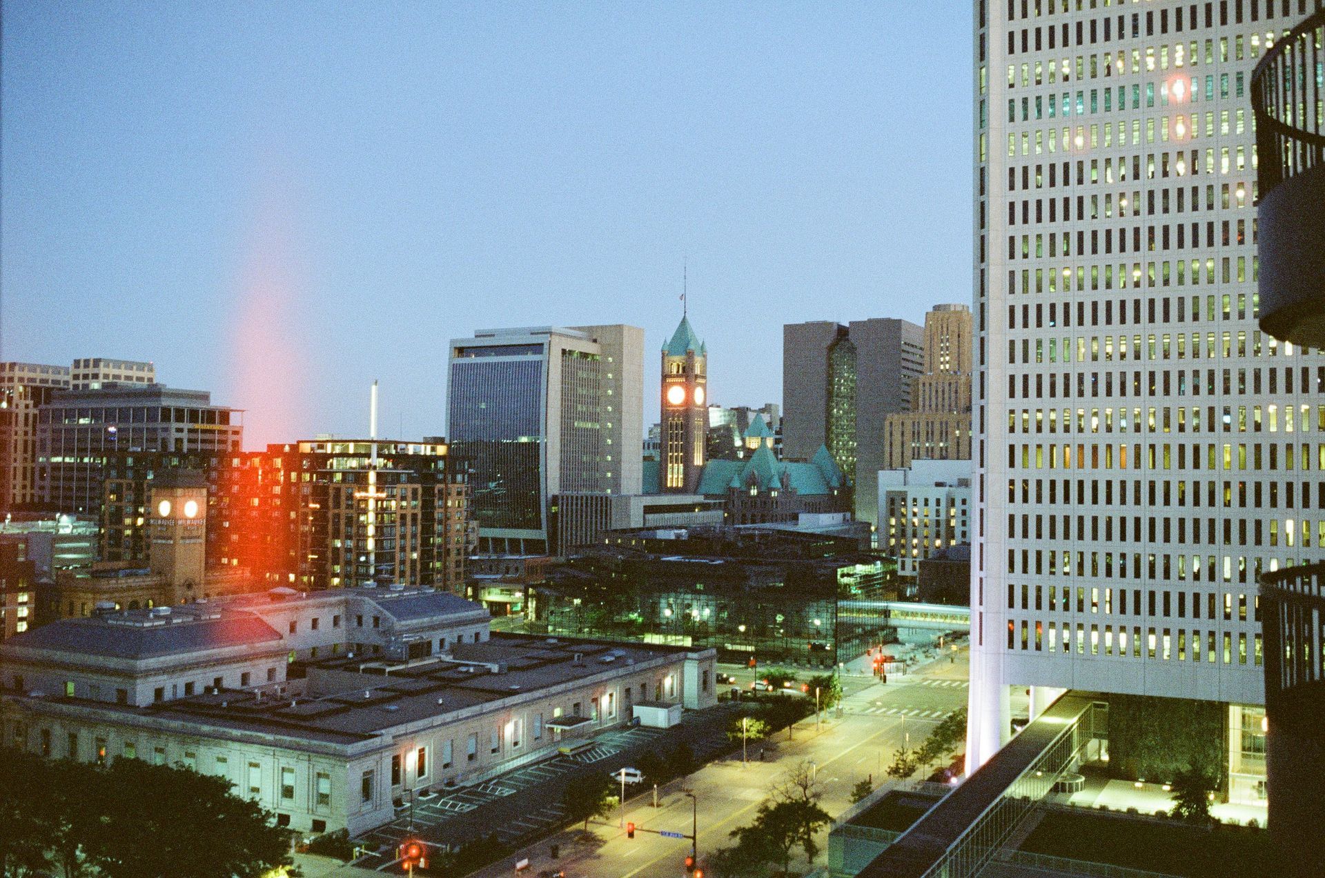 View of a city skyline at dusk, featuring a tall modern skyscraper, a historic government building, and glowing lights.