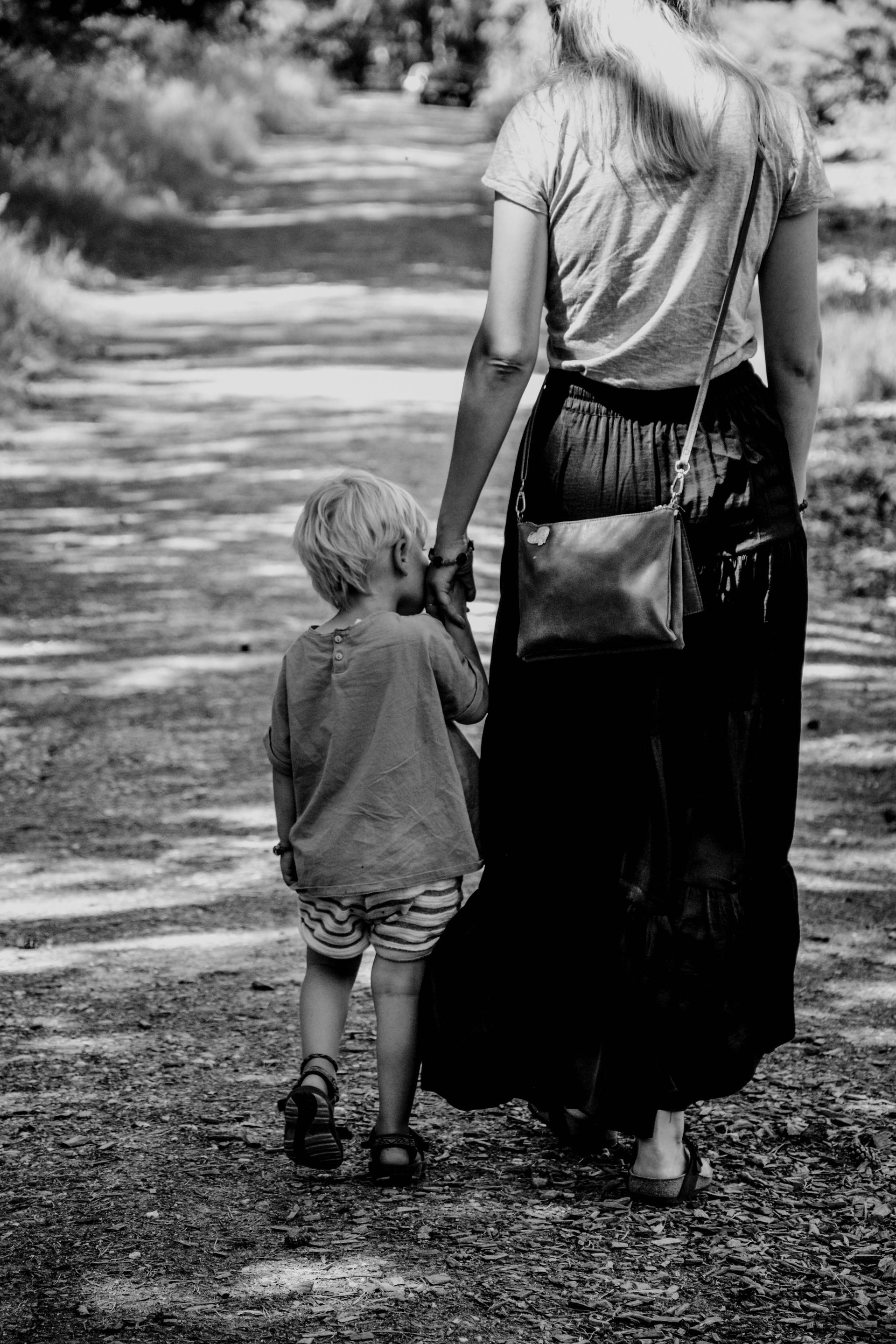 A person holds a child's hand while walking together along a sunlit, gravel path.