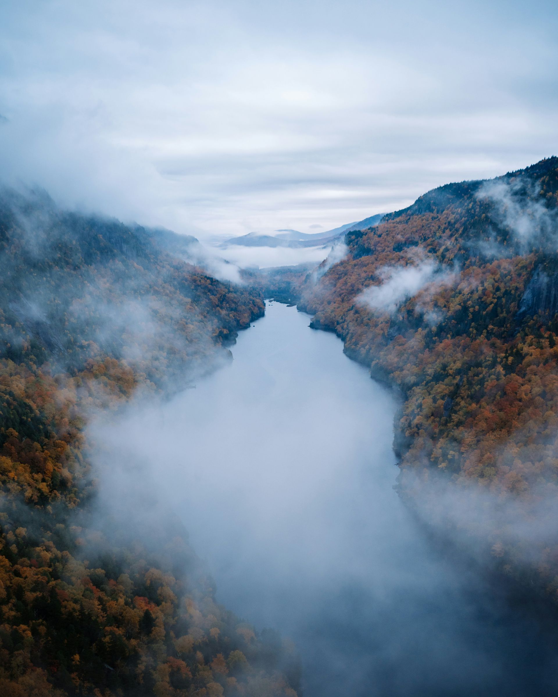 A winding river flows through a misty mountain valley surrounded by autumn-colored trees under a cloudy sky.