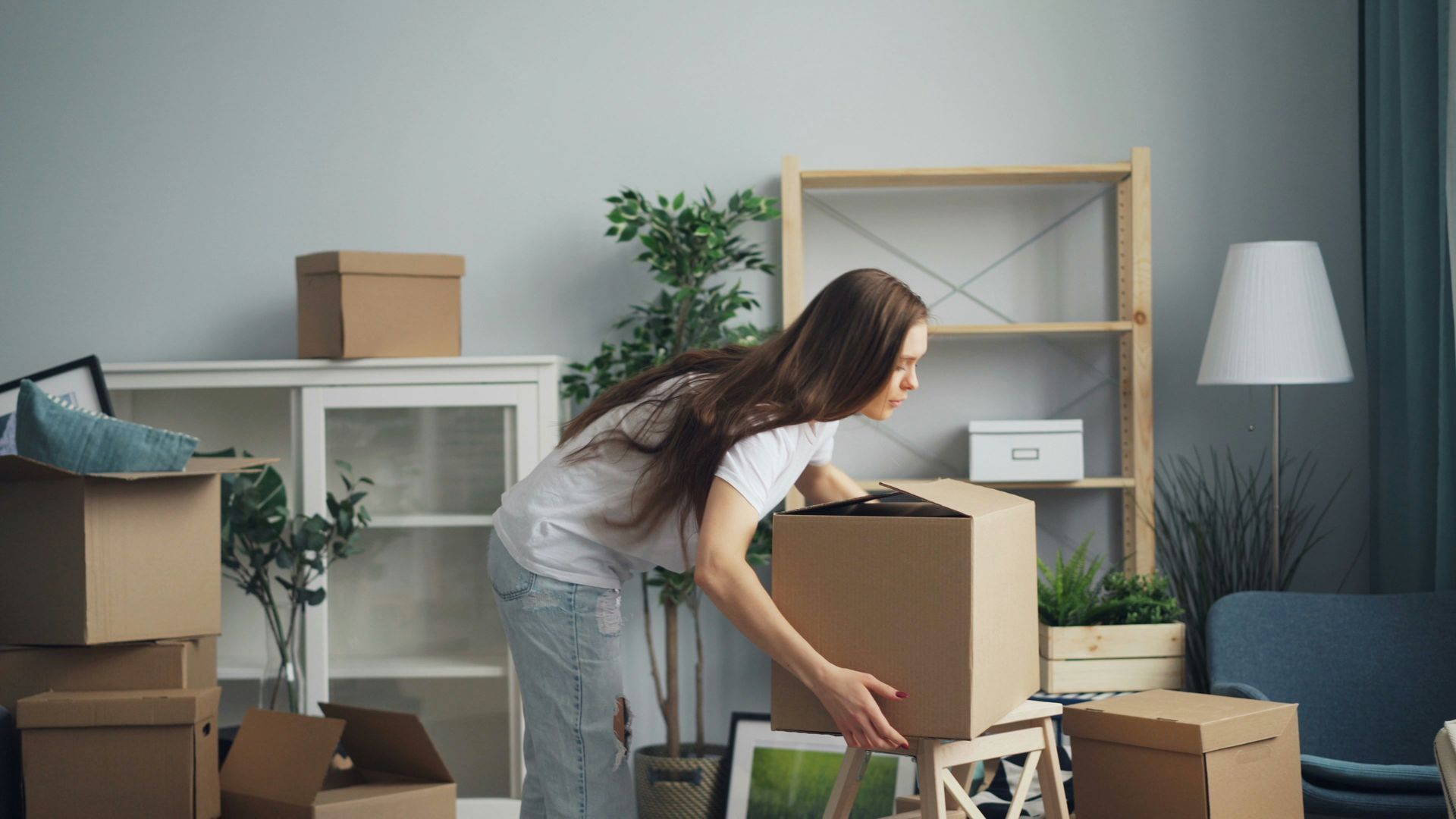 A person wearing a white shirt and jeans moves cardboard boxes in a room with a wooden shelf and a floor lamp.