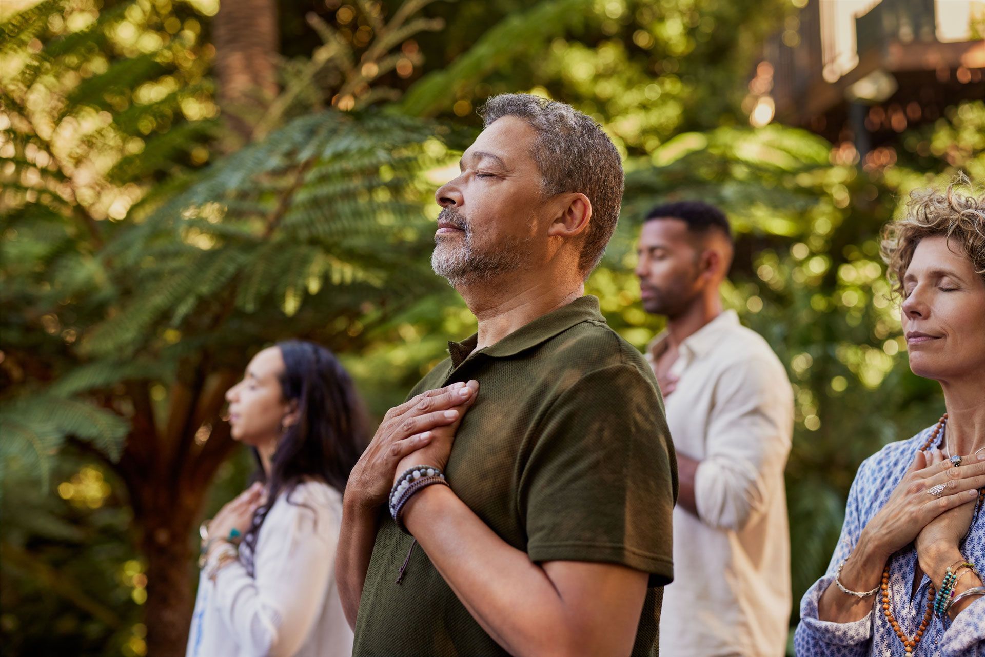 Four people in nature, meditating with hands on their chests, eyes closed. Green foliage in background.
