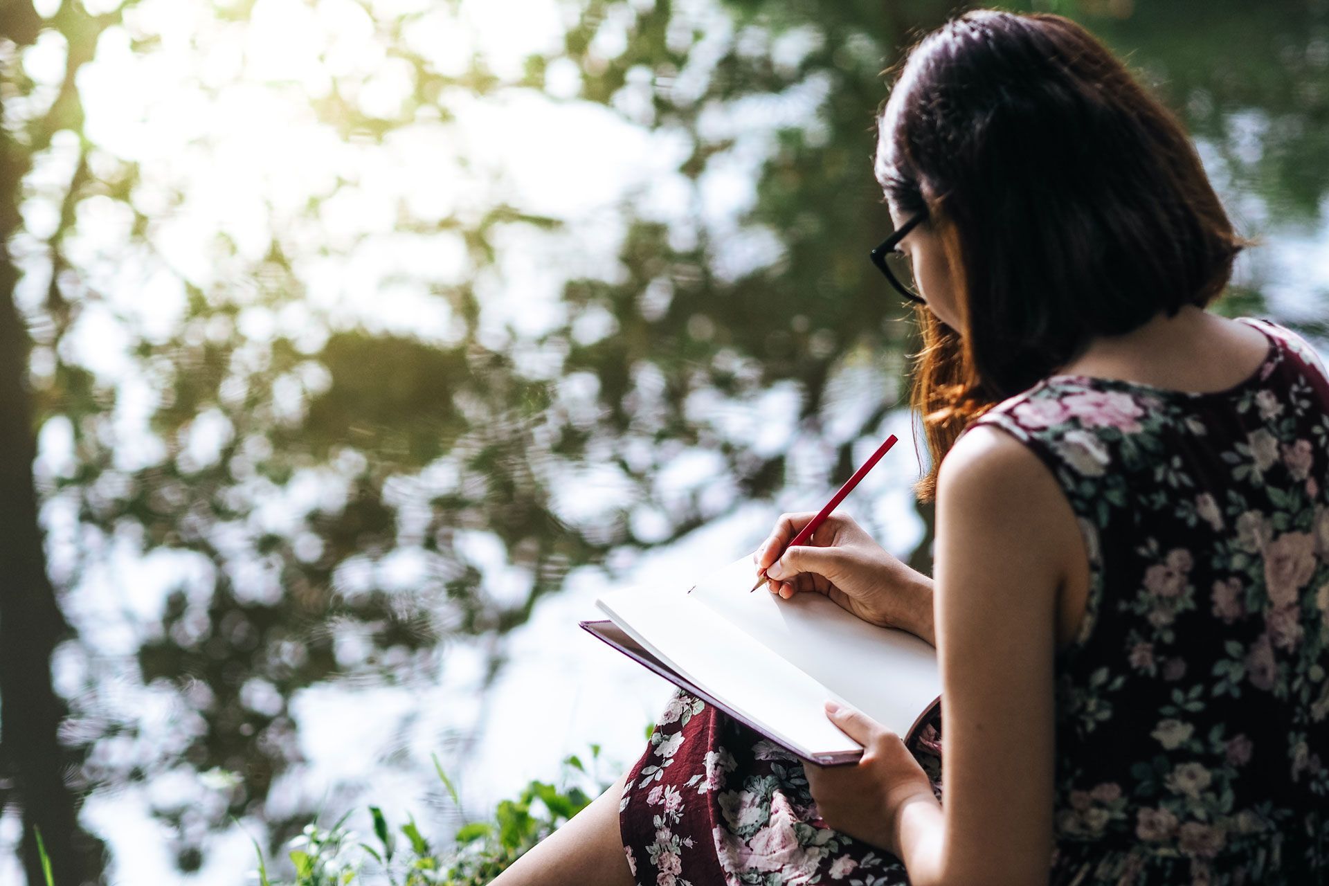 Woman with glasses, writing in notebook outdoors with trees in the background, holding a red pencil.