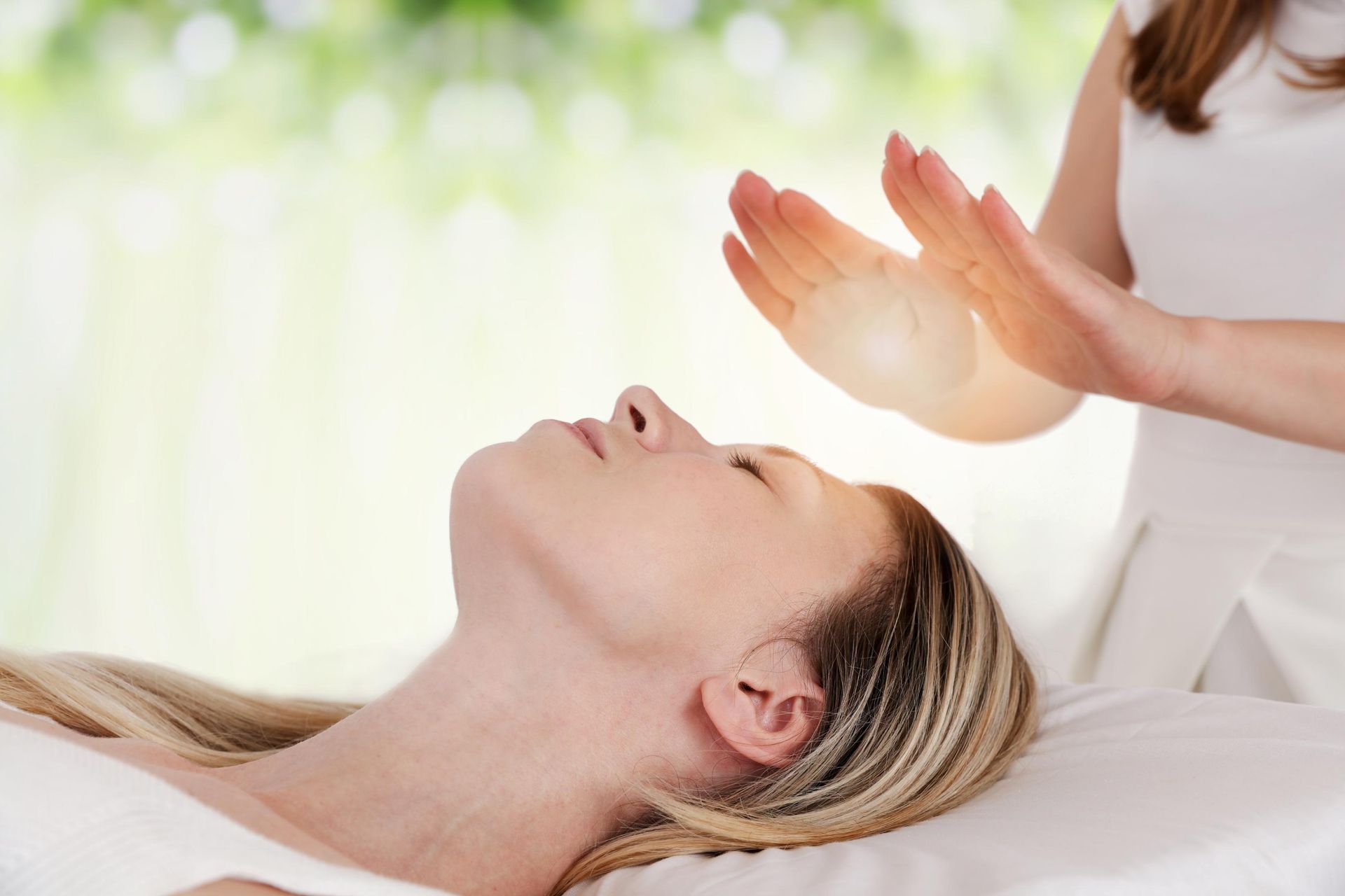 A practitioner holds their hands over a person's head, with a soft light glowing between their palms in a bright room.