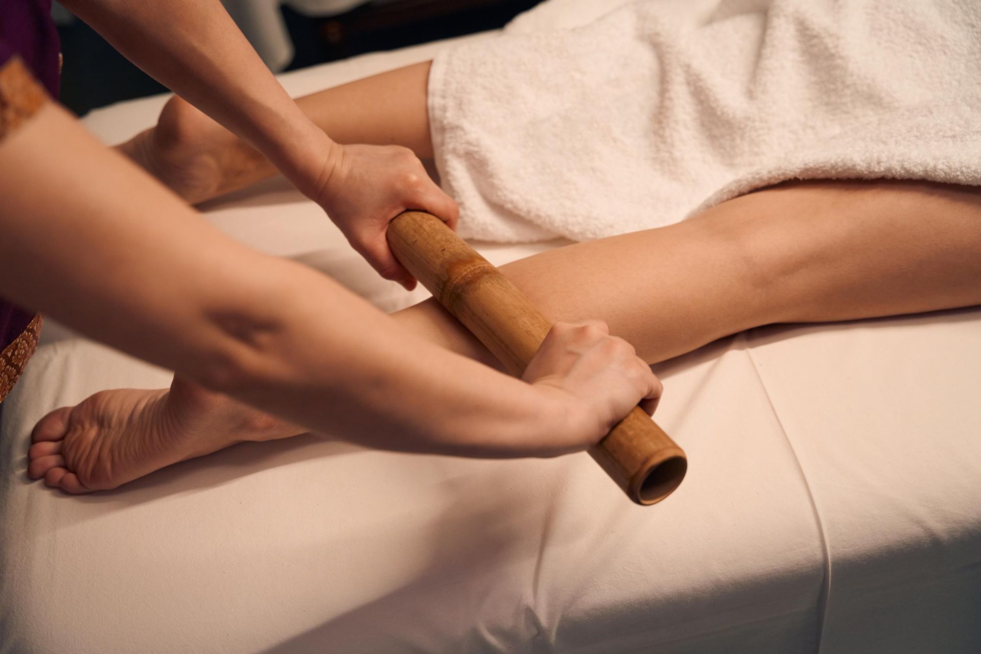 A person uses a bamboo roller tool to give a leg massage on a white massage table.