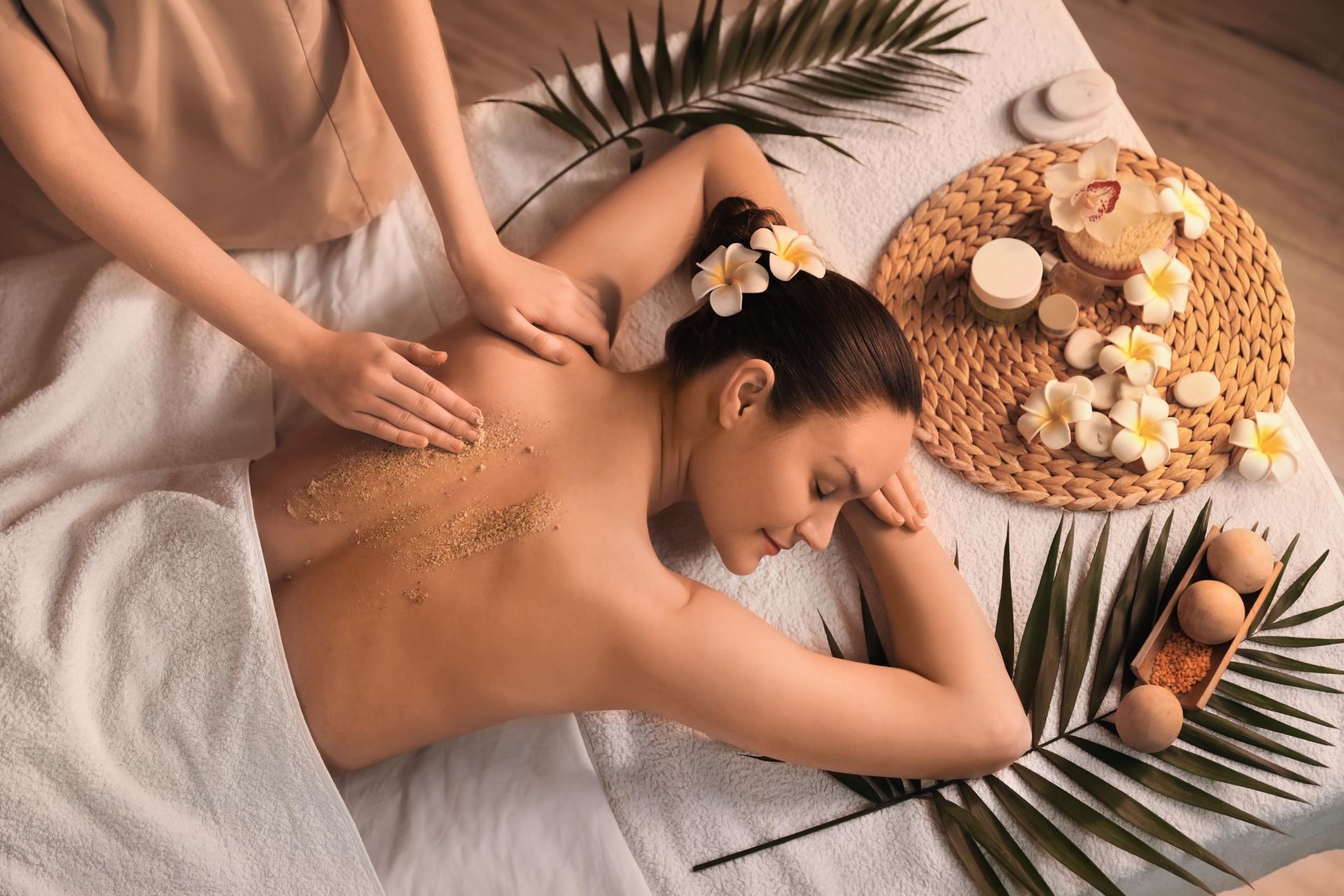 A massage therapist applies a scrub to a person’s back on a spa table decorated with tropical flowers and palm leaves.