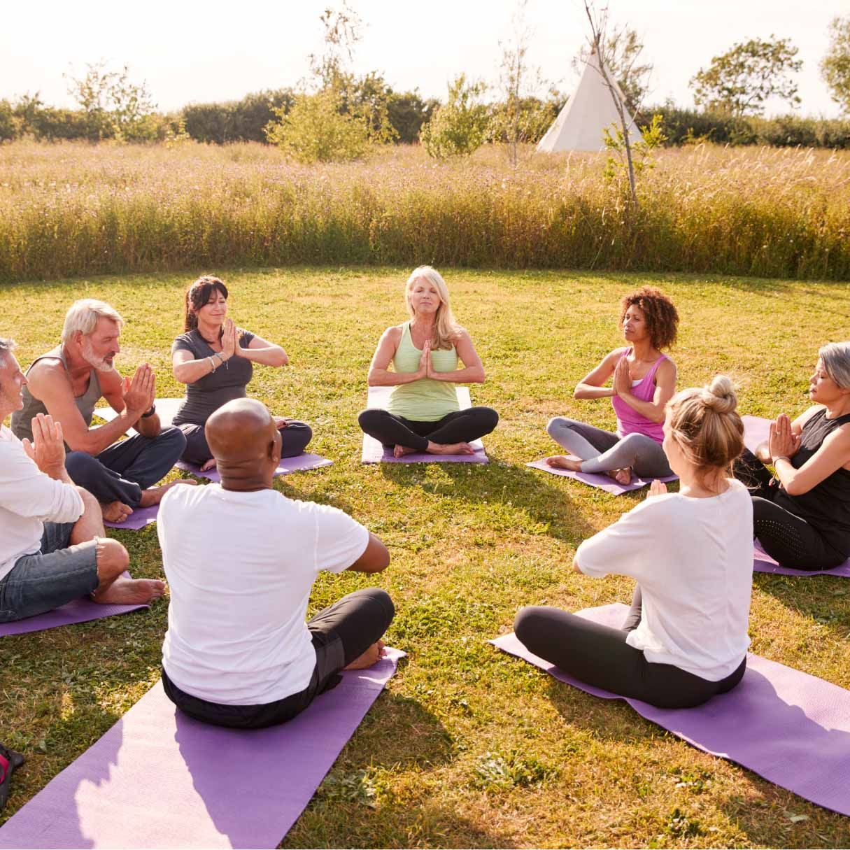 Eine Gruppe von Menschen in einem Kreis macht im Freien auf Matten Yoga, die Hände in Gebetshaltung, auf einem sonnigen Feld.