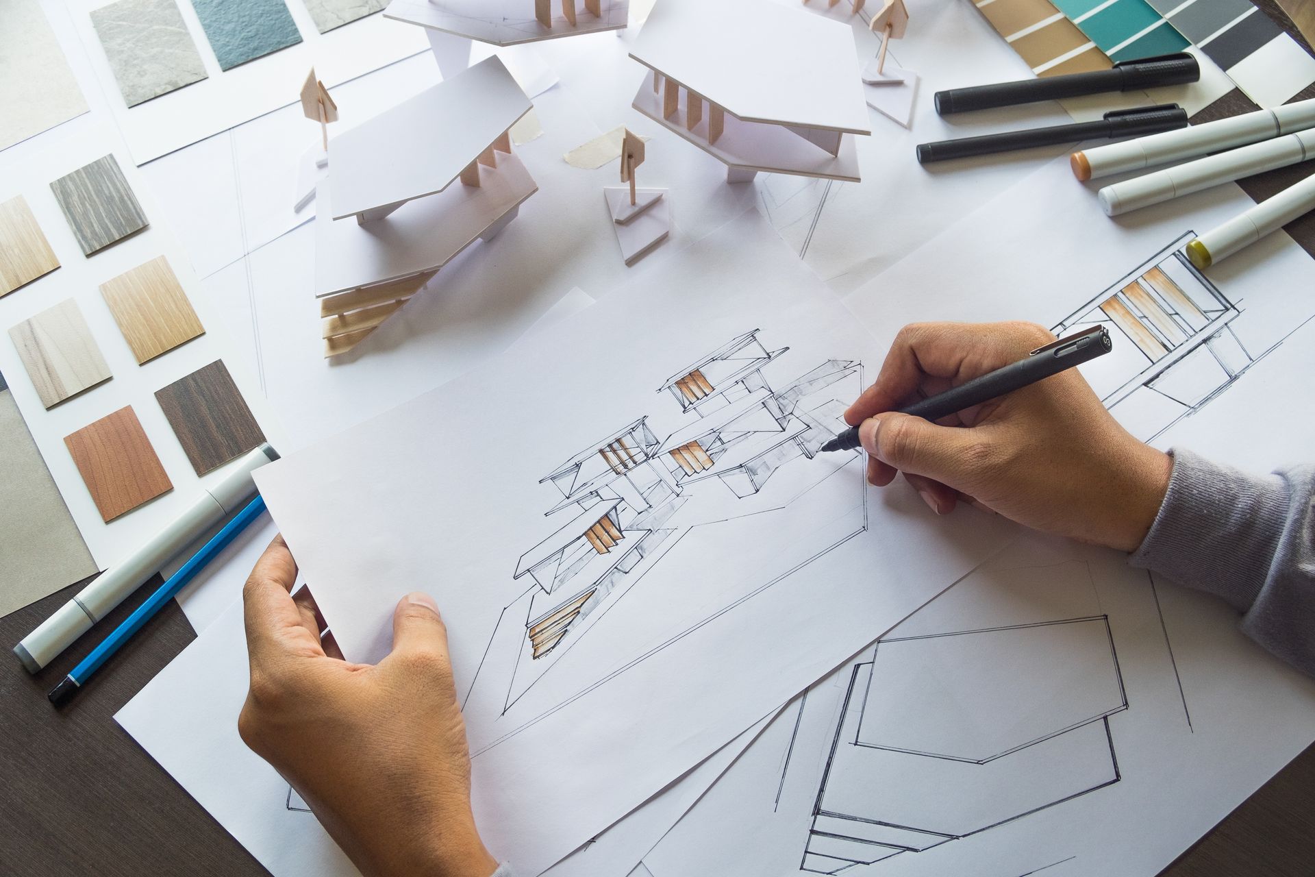 Hands sketching a house floor plan with pencils, architectural models, and color swatches on a desk