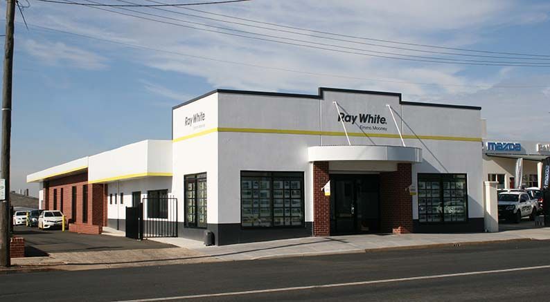 Street view of a white commercial building with red trim and storefront windows on a sunny day