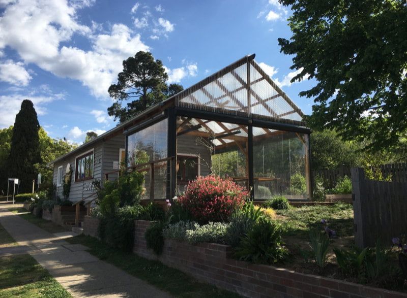 Glass greenhouse attached to a house, surrounded by a garden under a blue sky.