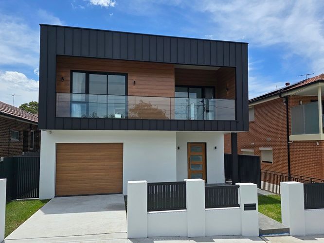 Modern two-story house with black and wood facade, balcony, garage, and white front fence