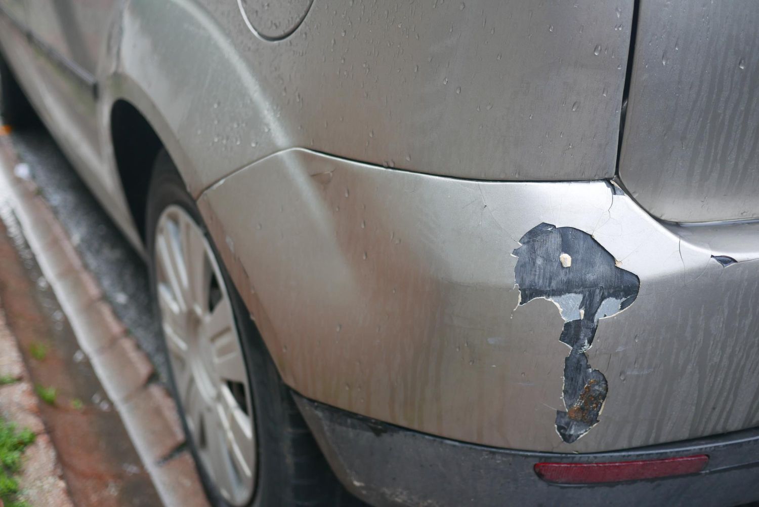 Close-up of a silver car bumper with chipped black paint damage near the rear wheel.