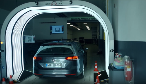 A grey station wagon car drives through a lit tunnel scanner in a workshop.